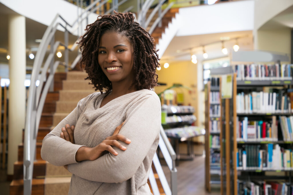 Woman standing with her arms crossed in a library, smiling near bookshelves and a staircase.