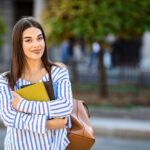 Student carrying books and a shoulder bag, standing on a city street with trees in the background.