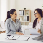 Two women sitting at a desk in an office, smiling and reviewing documents together.