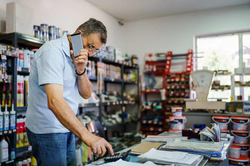 Store owner using a calculator while talking on a phone behind a shop counter with paperwork