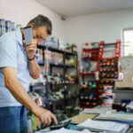 Store owner using a calculator while talking on a phone behind a shop counter with paperwork