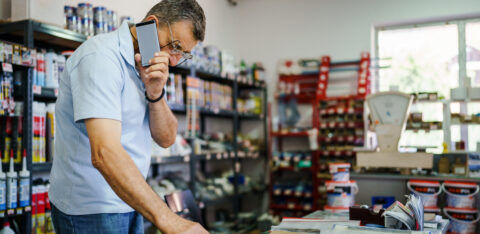 Store owner using a calculator while talking on a phone behind a shop counter with paperwork