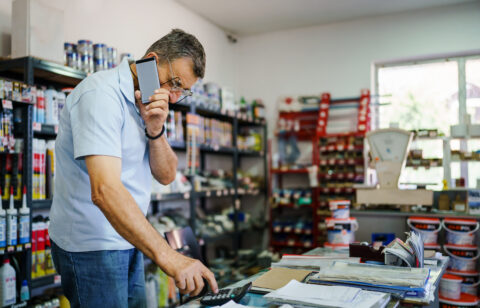 Store owner using a calculator while talking on a phone behind a shop counter with paperwork
