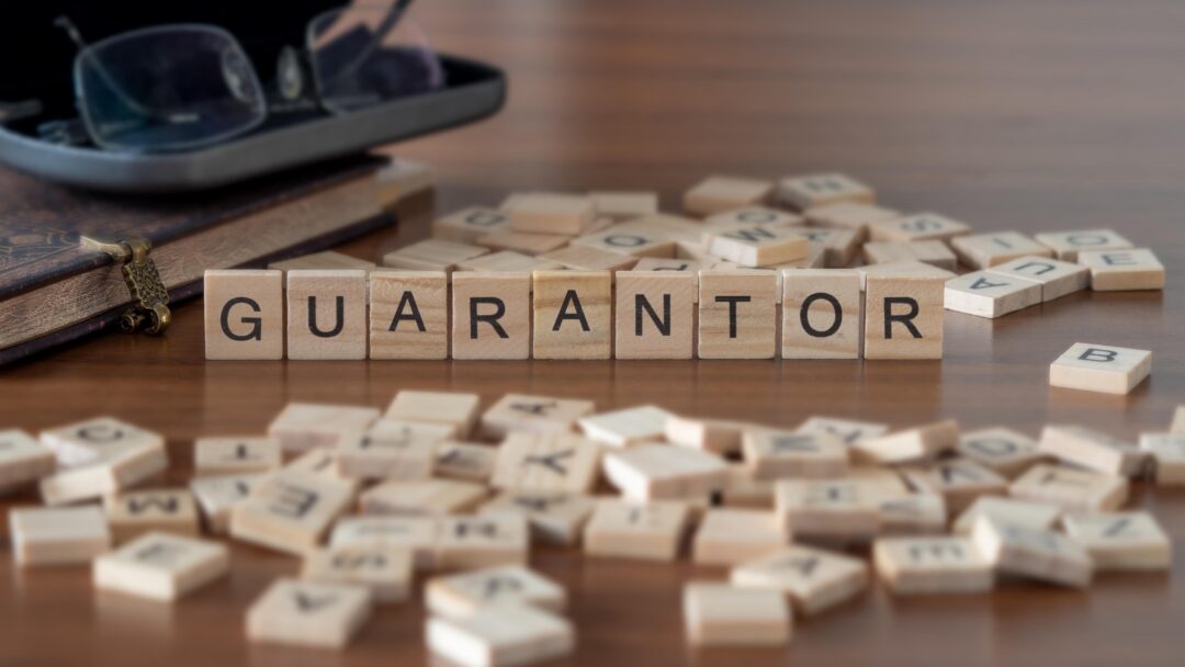 Wooden letter tiles spelling “guarantor” on a table, with scattered tiles and books in the background.