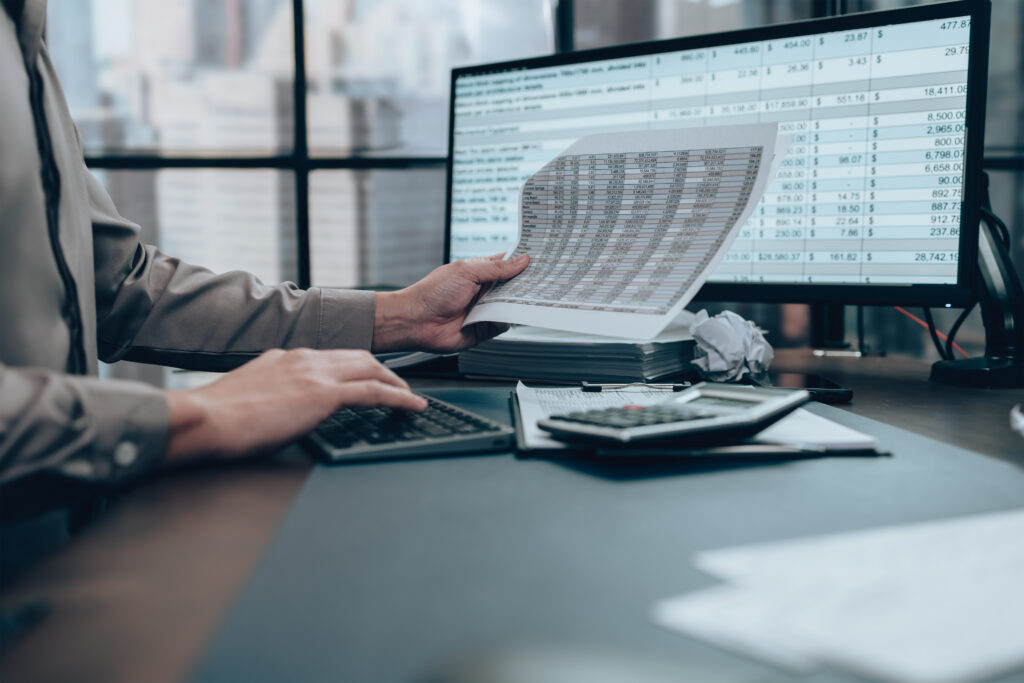 Person reviewing financial documents while using a calculator and computer with spreadsheet data on screen