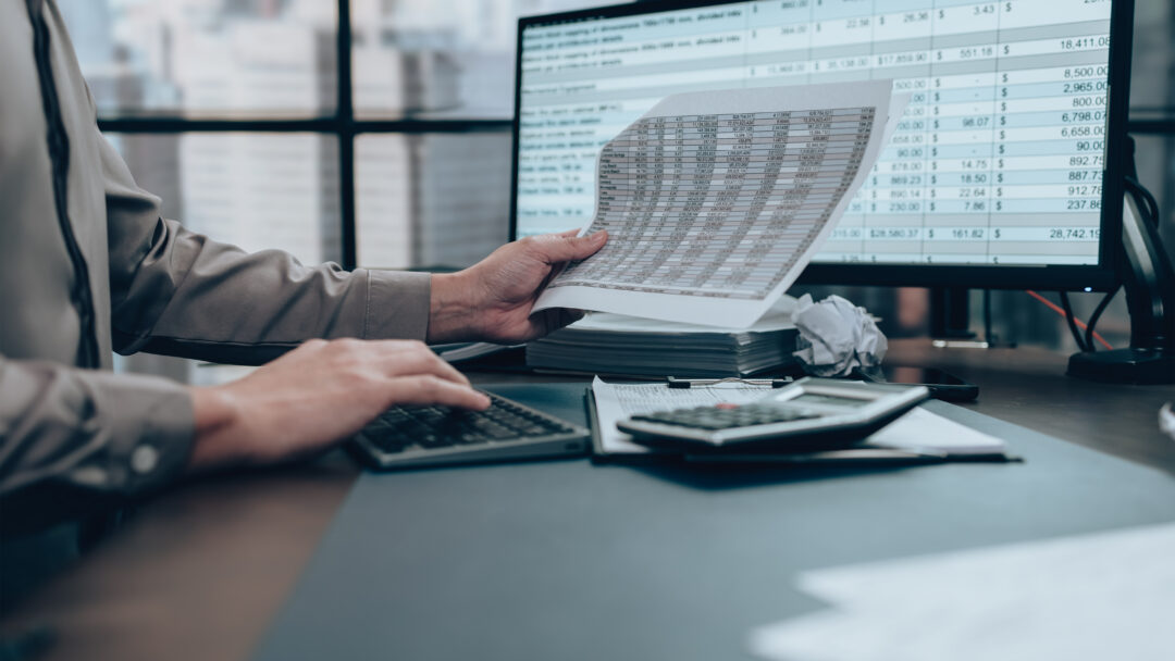 Person reviewing financial documents while using a calculator and computer with spreadsheet data on screen