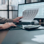 Person reviewing financial documents while using a calculator and computer with spreadsheet data on screen