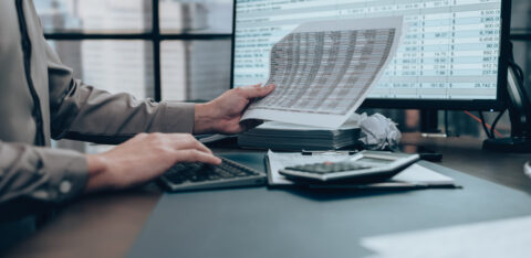Person reviewing financial documents while using a calculator and computer with spreadsheet data on screen