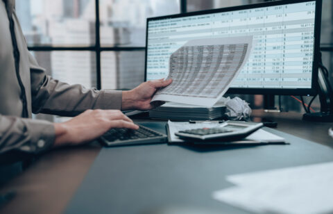 Person reviewing financial documents while using a calculator and computer with spreadsheet data on screen