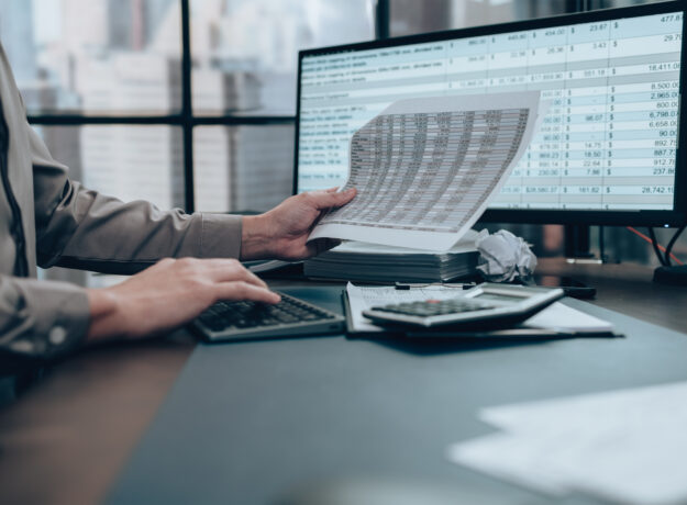 Person reviewing financial documents while using a calculator and computer with spreadsheet data on screen
