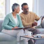 Older couple reviewing paperwork together on a couch, with one holding a document and the other taking notes