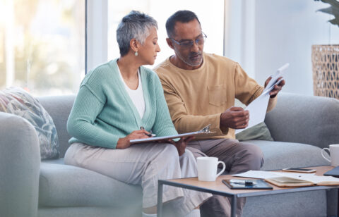 Older couple reviewing paperwork together on a couch, with one holding a document and the other taking notes