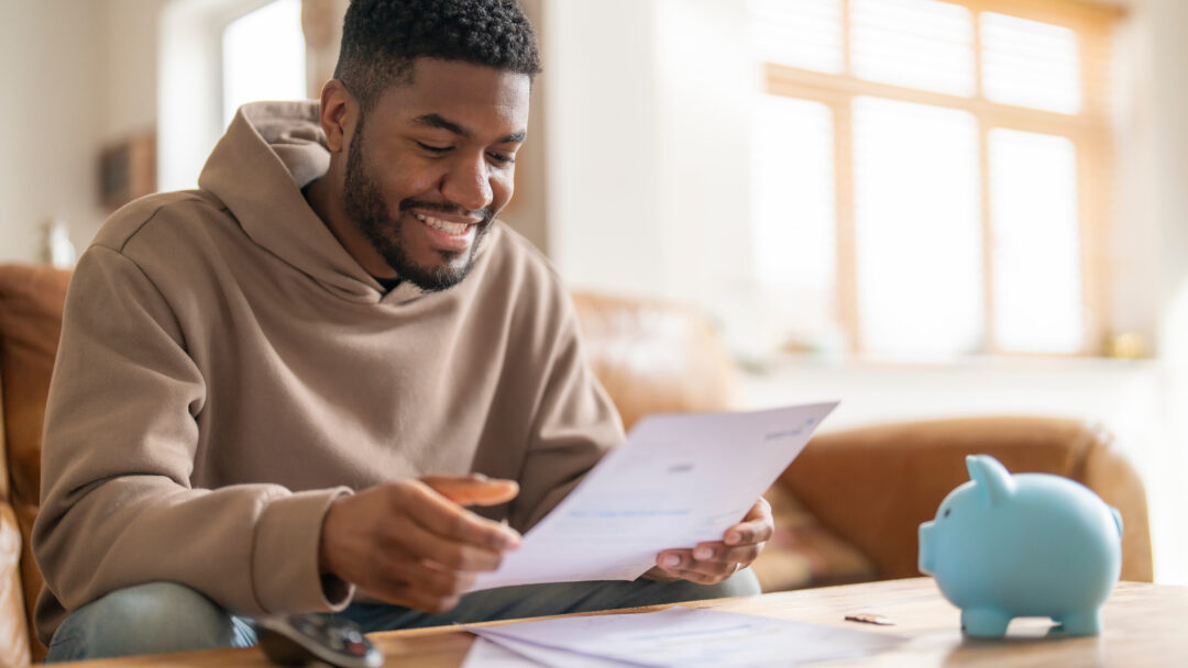 Person sitting at a table smiling while reviewing a document, with a piggy bank nearby