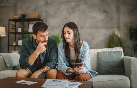 Two adults looking at financial documents together at home, checking information on a smartphone.