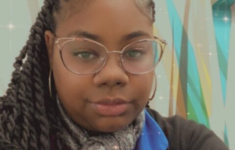 Woman with braided hair and glasses taking a selfie indoors, wearing a scarf and blue shirt