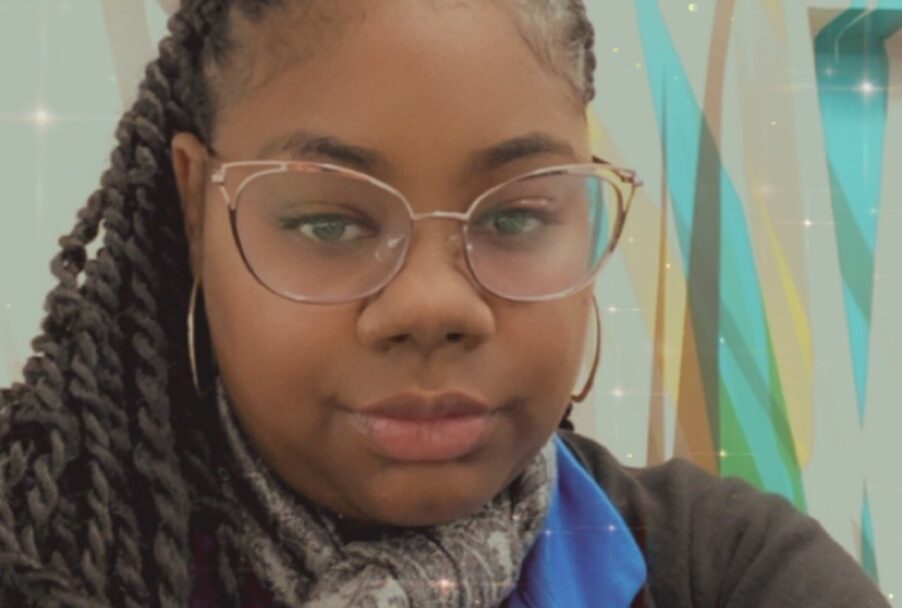 Woman with braided hair and glasses taking a selfie indoors, wearing a scarf and blue shirt