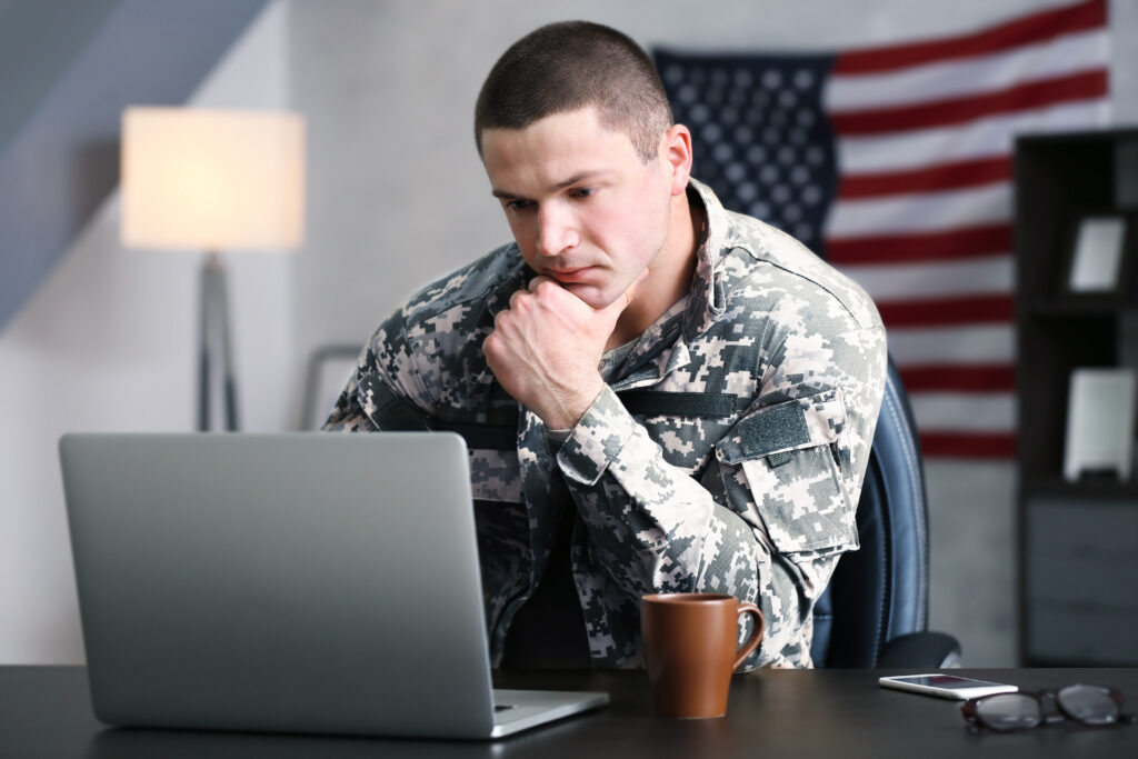 Person in military uniform sitting at a desk, looking at a laptop with a thoughtful expression, American flag in the background