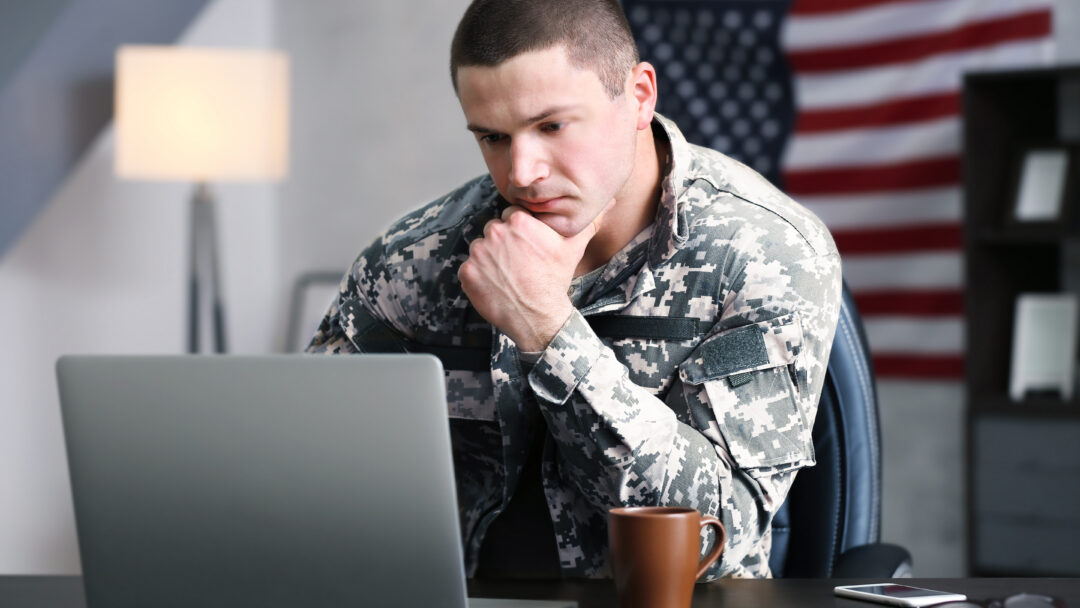 Person in military uniform sitting at a desk, looking at a laptop with a thoughtful expression, American flag in the background