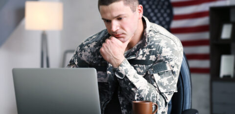 Person in military uniform sitting at a desk, looking at a laptop with a thoughtful expression, American flag in the background