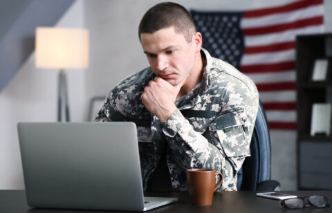 Person in military uniform sitting at a desk, looking at a laptop with a thoughtful expression, American flag in the background
