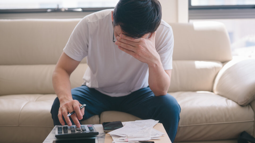 Person sitting on a couch reviewing bills, using a calculator while covering their face in stress