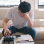 Person sitting on a couch reviewing bills, using a calculator while covering their face in stress
