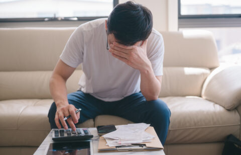 Person sitting on a couch reviewing bills, using a calculator while covering their face in stress