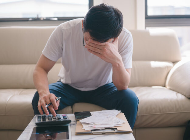 Person sitting on a couch reviewing bills, using a calculator while covering their face in stress