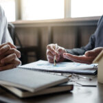 Two people reviewing financial documents at a desk, with one person pointing to a chart on a report.
