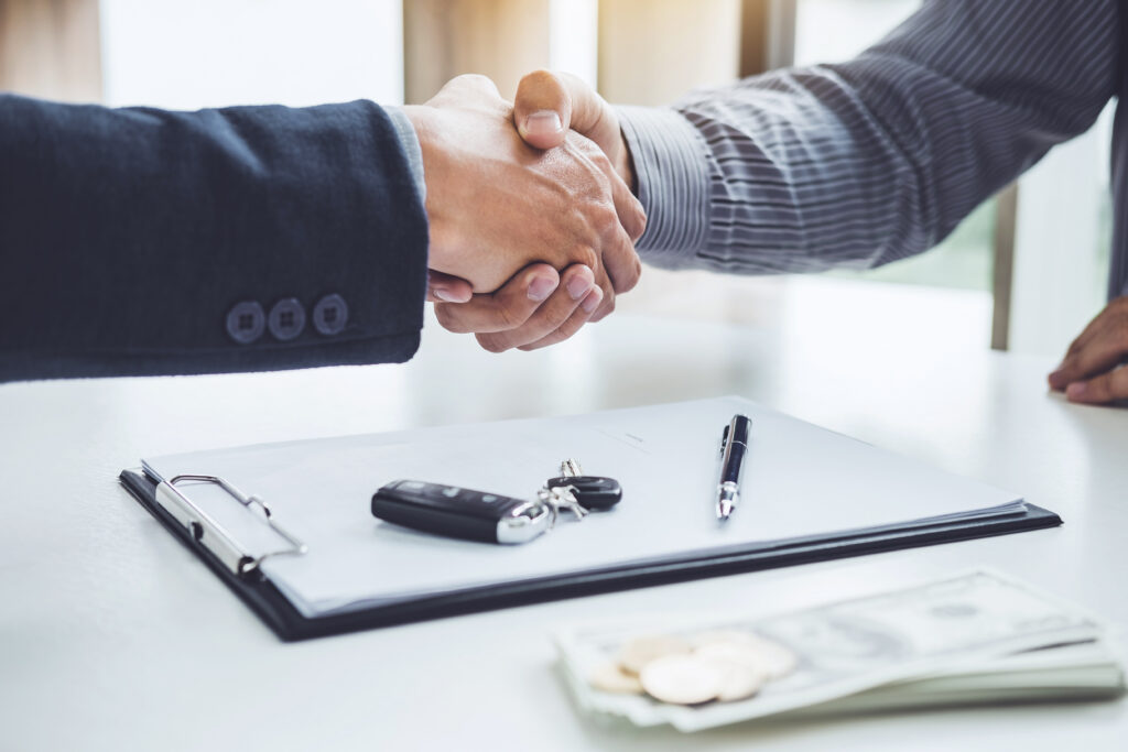 Two people shaking hands across a desk with a clipboard, car keys, pen, and cash visible in the foreground.