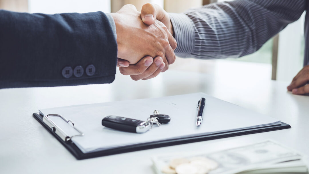 Two people shaking hands across a desk with a clipboard, car keys, pen, and cash visible in the foreground.