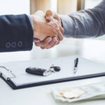 Two people shaking hands across a desk with a clipboard, car keys, pen, and cash visible in the foreground.