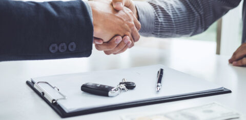 Two people shaking hands across a desk with a clipboard, car keys, pen, and cash visible in the foreground.