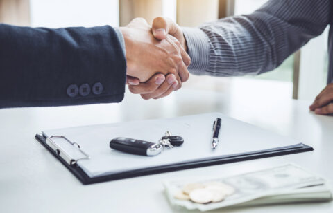 Two people shaking hands across a desk with a clipboard, car keys, pen, and cash visible in the foreground.