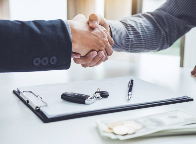 Two people shaking hands across a desk with a clipboard, car keys, pen, and cash visible in the foreground.