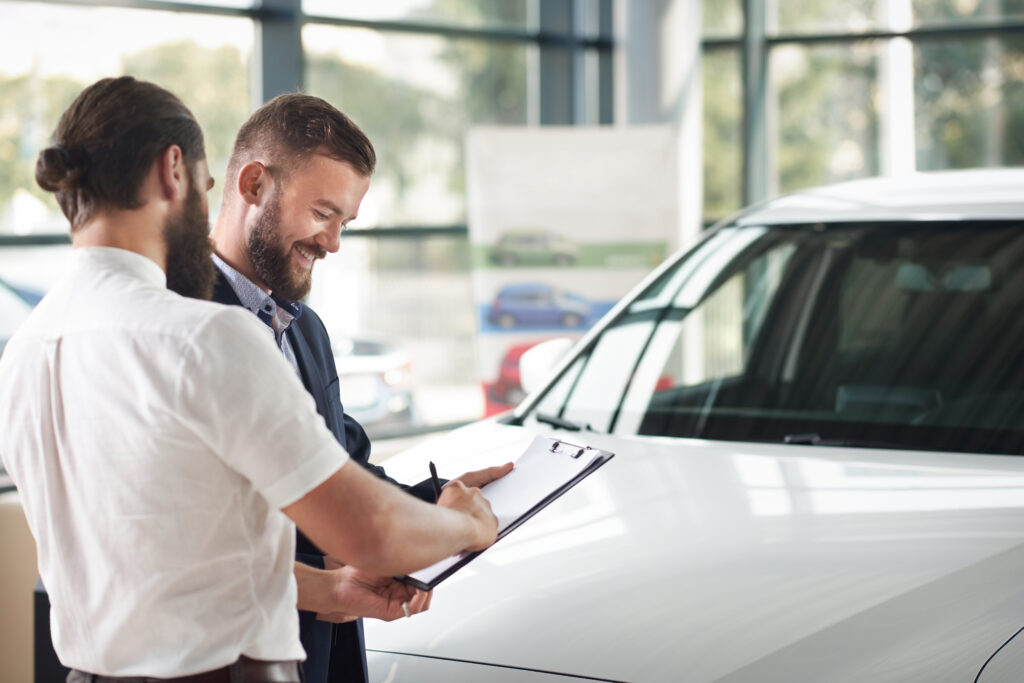 Customer signing paperwork on a clipboard while standing next to a car with a salesperson