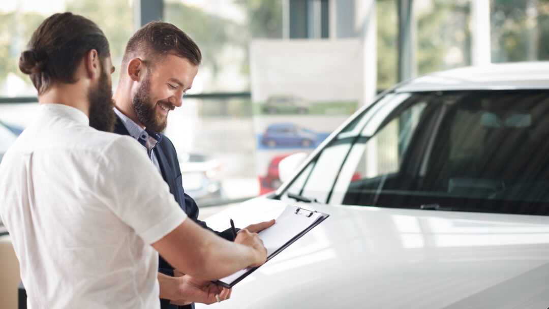 Customer signing paperwork on a clipboard while standing next to a car with a salesperson