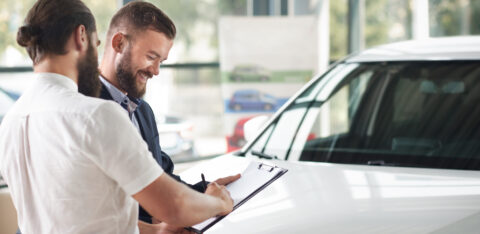Customer signing paperwork on a clipboard while standing next to a car with a salesperson