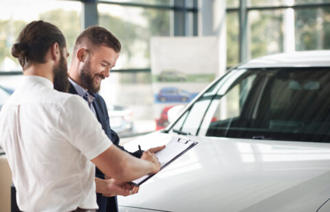 Customer signing paperwork on a clipboard while standing next to a car with a salesperson