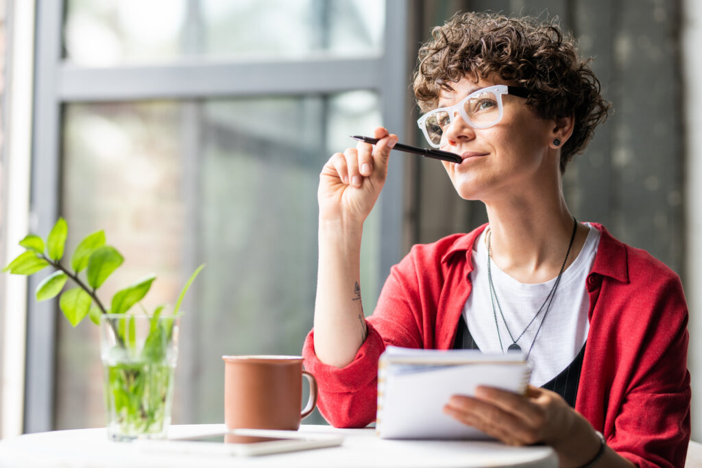 Woman sitting by a window, holding a notebook and pen while looking thoughtfully into the distance.