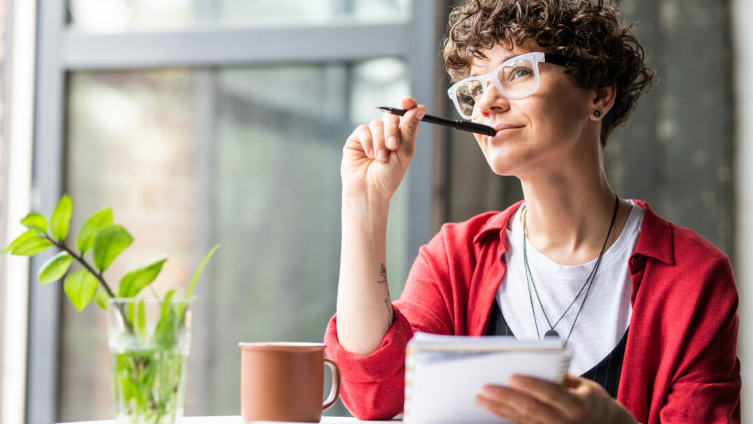 Woman sitting by a window, holding a notebook and pen while looking thoughtfully into the distance.