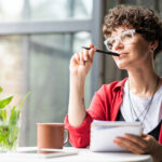 Woman sitting by a window, holding a notebook and pen while looking thoughtfully into the distance.