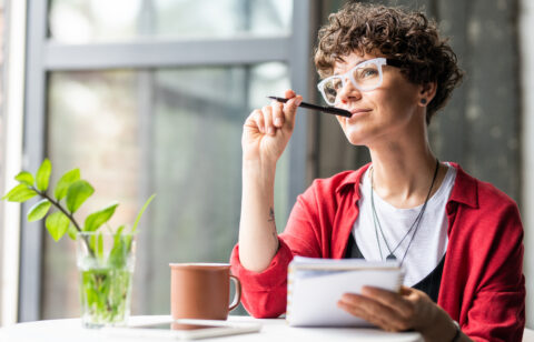 Woman sitting by a window, holding a notebook and pen while looking thoughtfully into the distance.