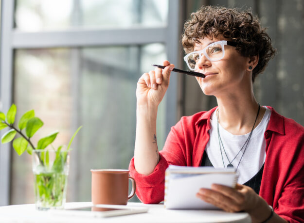 Woman sitting by a window, holding a notebook and pen while looking thoughtfully into the distance.