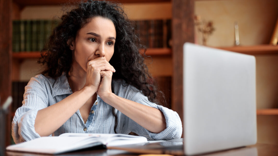 Woman sitting at a desk and looking thoughtfully at a laptop, with an open notebook in front of her.