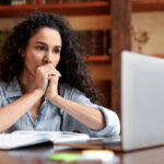Woman sitting at a desk and looking thoughtfully at a laptop, with an open notebook in front of her.