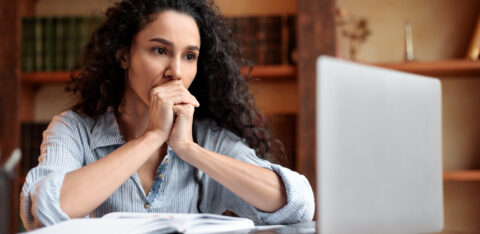 Woman sitting at a desk and looking thoughtfully at a laptop, with an open notebook in front of her.