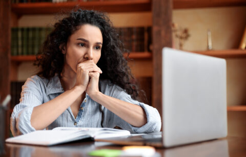 Woman sitting at a desk and looking thoughtfully at a laptop, with an open notebook in front of her.