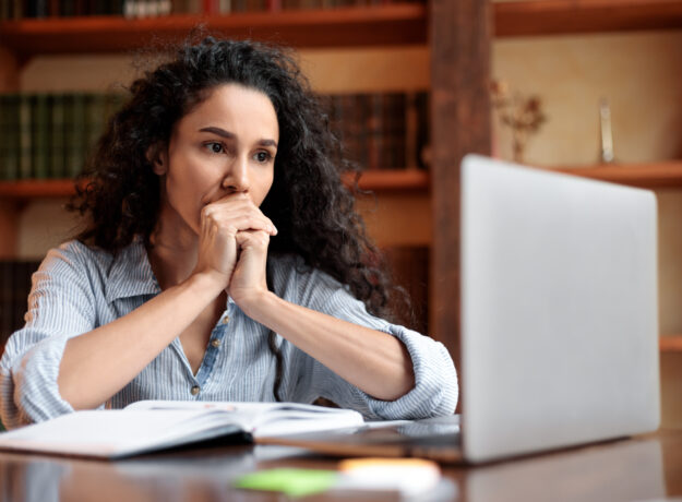 Woman sitting at a desk and looking thoughtfully at a laptop, with an open notebook in front of her.