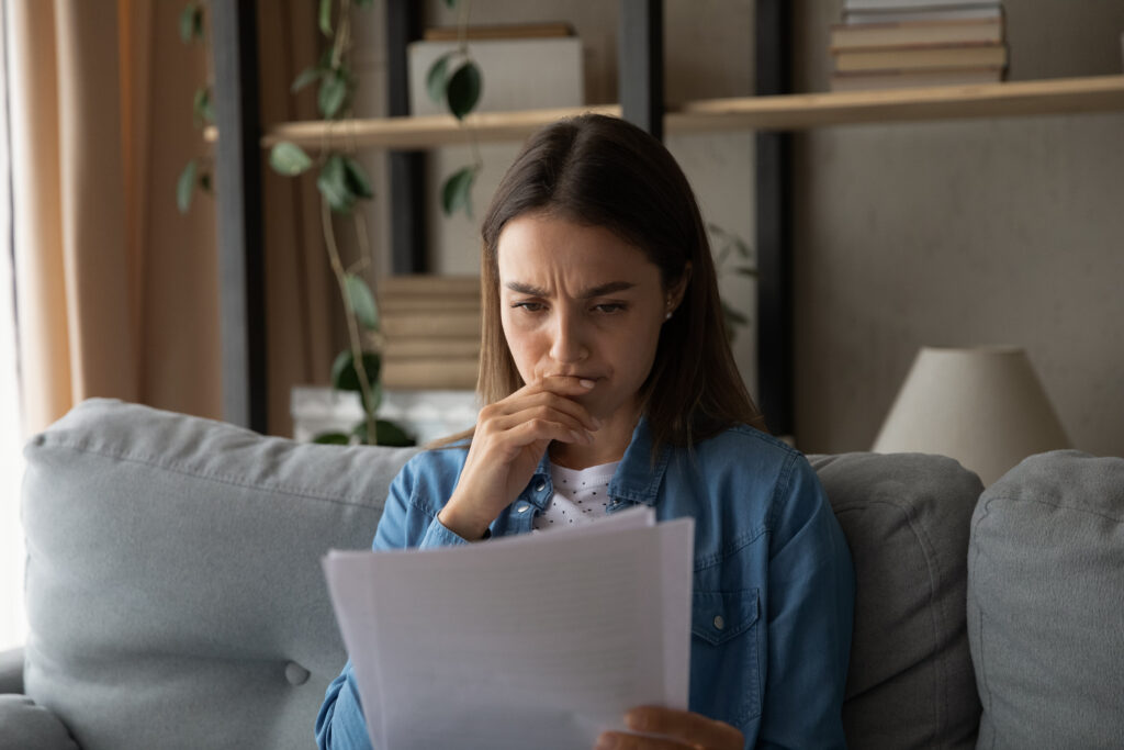 Person sitting on a couch reading a document with a concerned expression, hand near their mouth